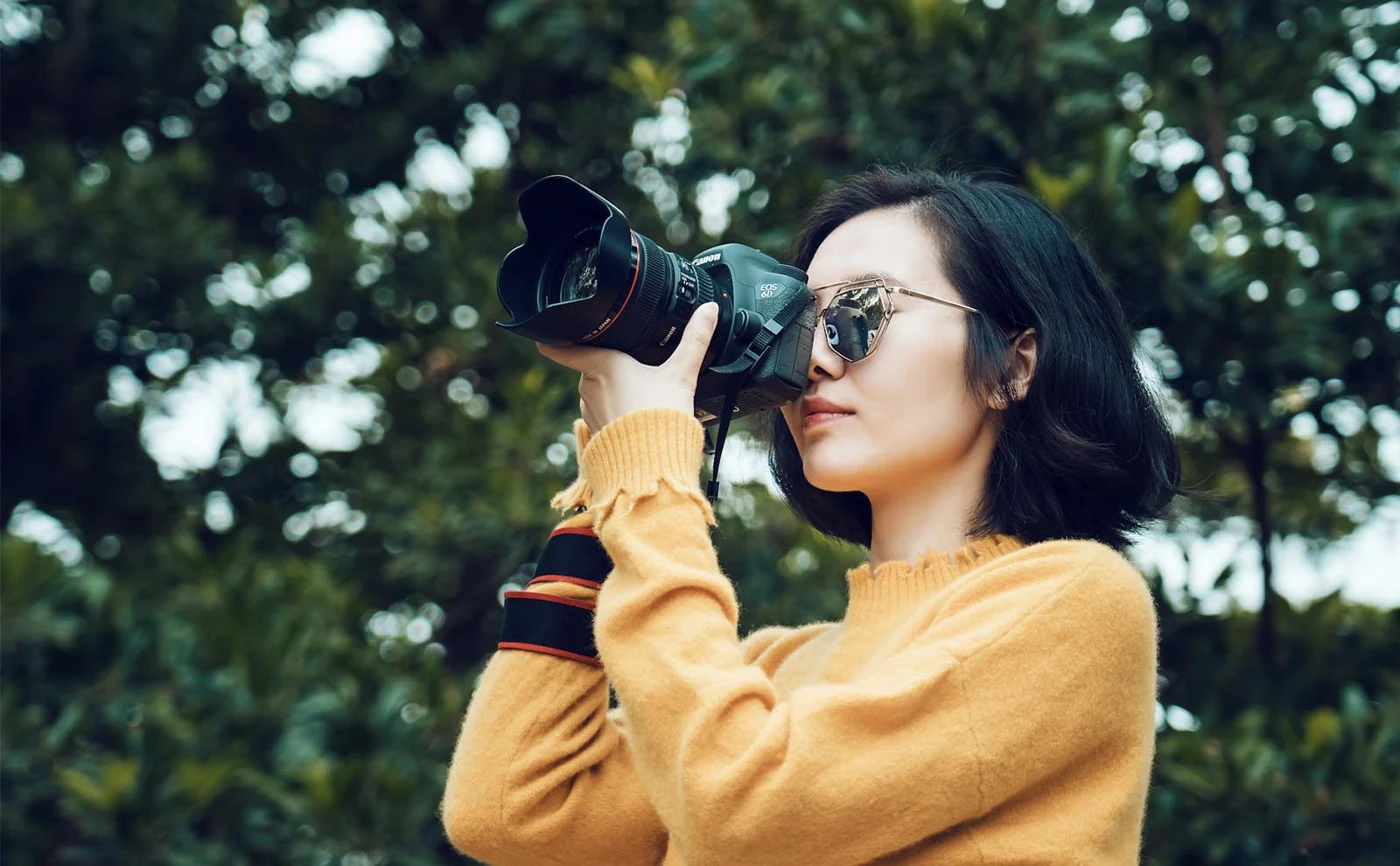 Woman in a mustard sweater wearing sunglasses and holding a camera outdoors.