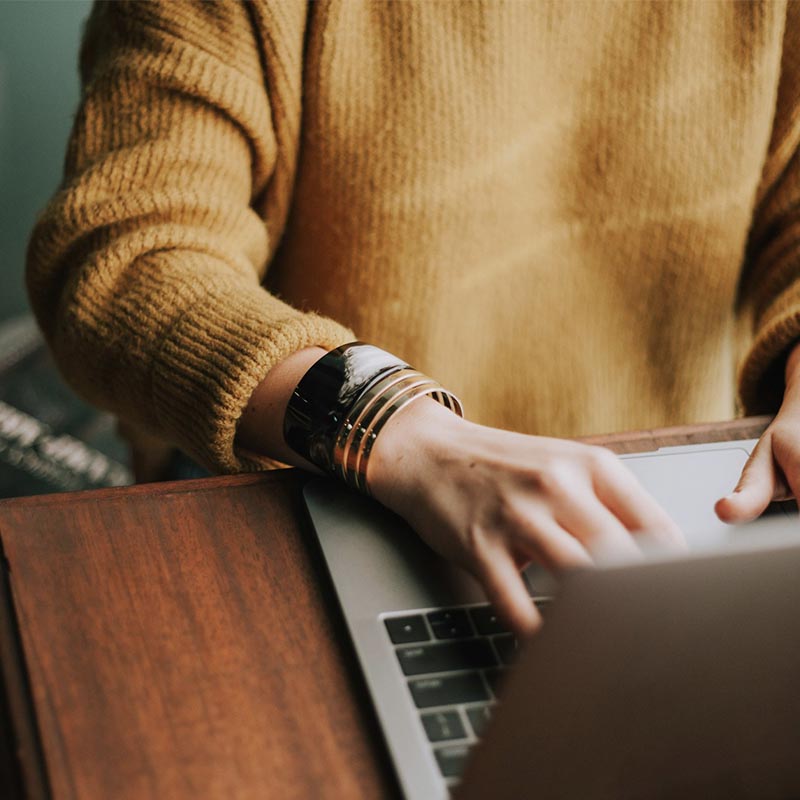 Close-up of a person in a mustard yellow sweater typing on a laptop, wearing a black and gold bracelet.