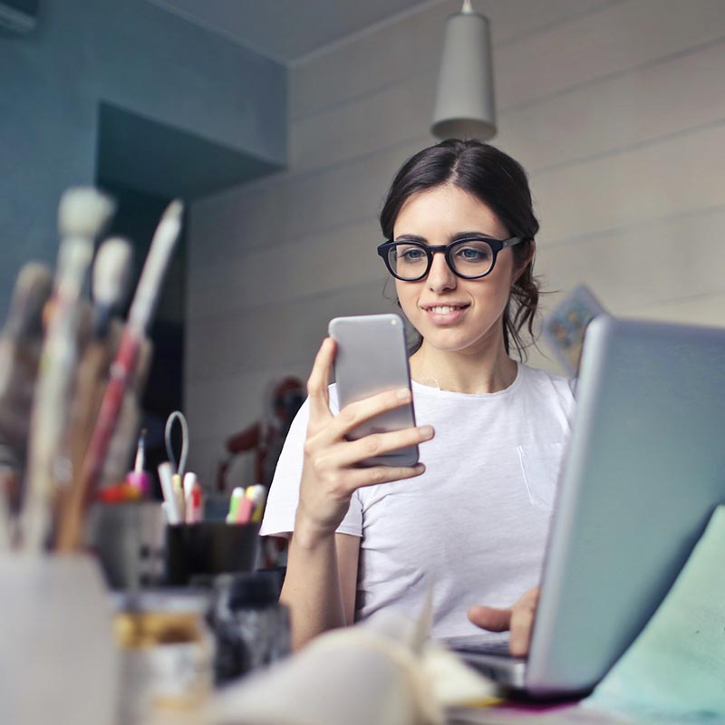 Woman wearing glasses, smiling while looking at her phone, with a laptop and art supplies on her desk.